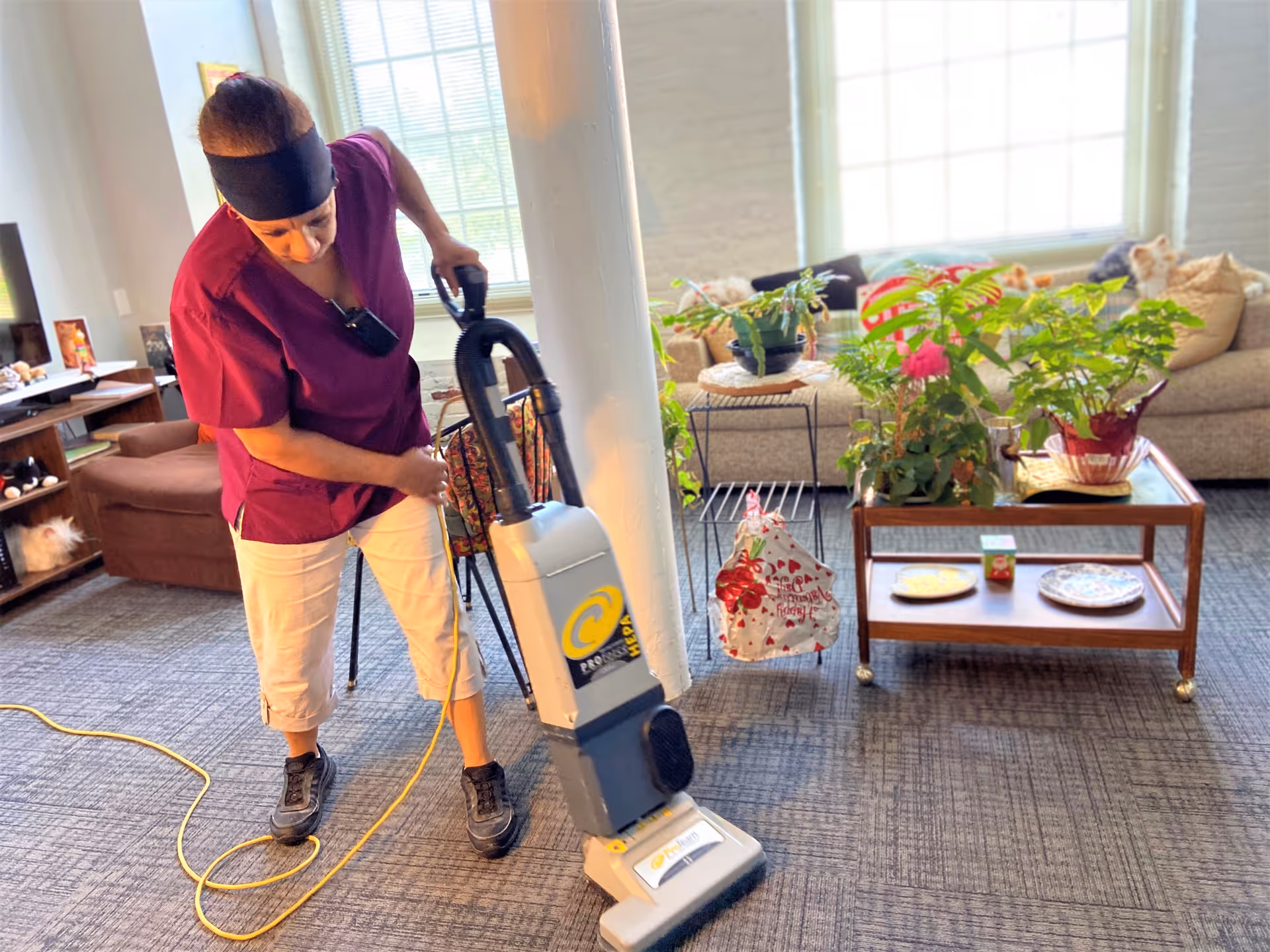 A person wearing a maroon top and white pants is vacuuming a carpeted floor in a living room area. The room has a beige couch with several cushions, a wooden coffee table with potted plants and decorative plates, and large windows letting in natural light.