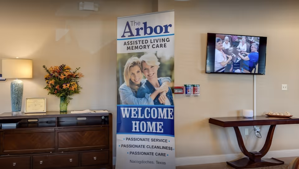 Welcoming lobby area with a tall "The Arbor" welcome banner, a dresser with a lamp and flowers, and a wall-mounted TV showing residents.