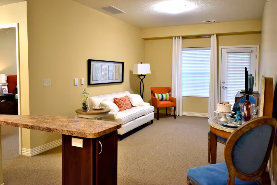A cozy living room area with beige walls and carpet. There is a white sofa with decorative pillows, a round side table with a glass jar and a plate, an orange armchair with a striped pillow, and a floor lamp. A small round dining table with two blue cushioned chairs is set with plates, glasses, and a cake. A window with white curtains and a door with blinds are visible in the background. A countertop with a granite finish extends into the room from the left side.