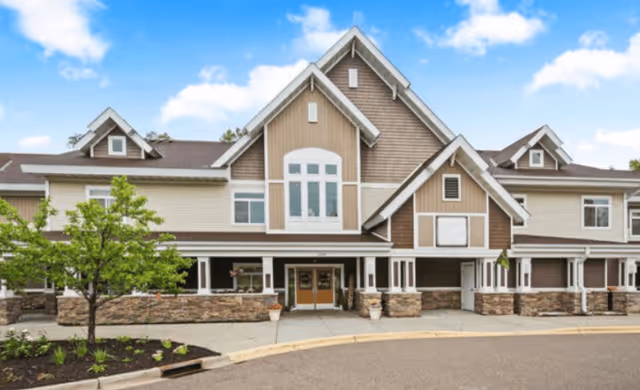 Front exterior view of a two-story assisted living and memory care facility with beige and brown siding, multiple gabled roofs, large windows, a covered entrance, and a small landscaped area with a tree and plants.