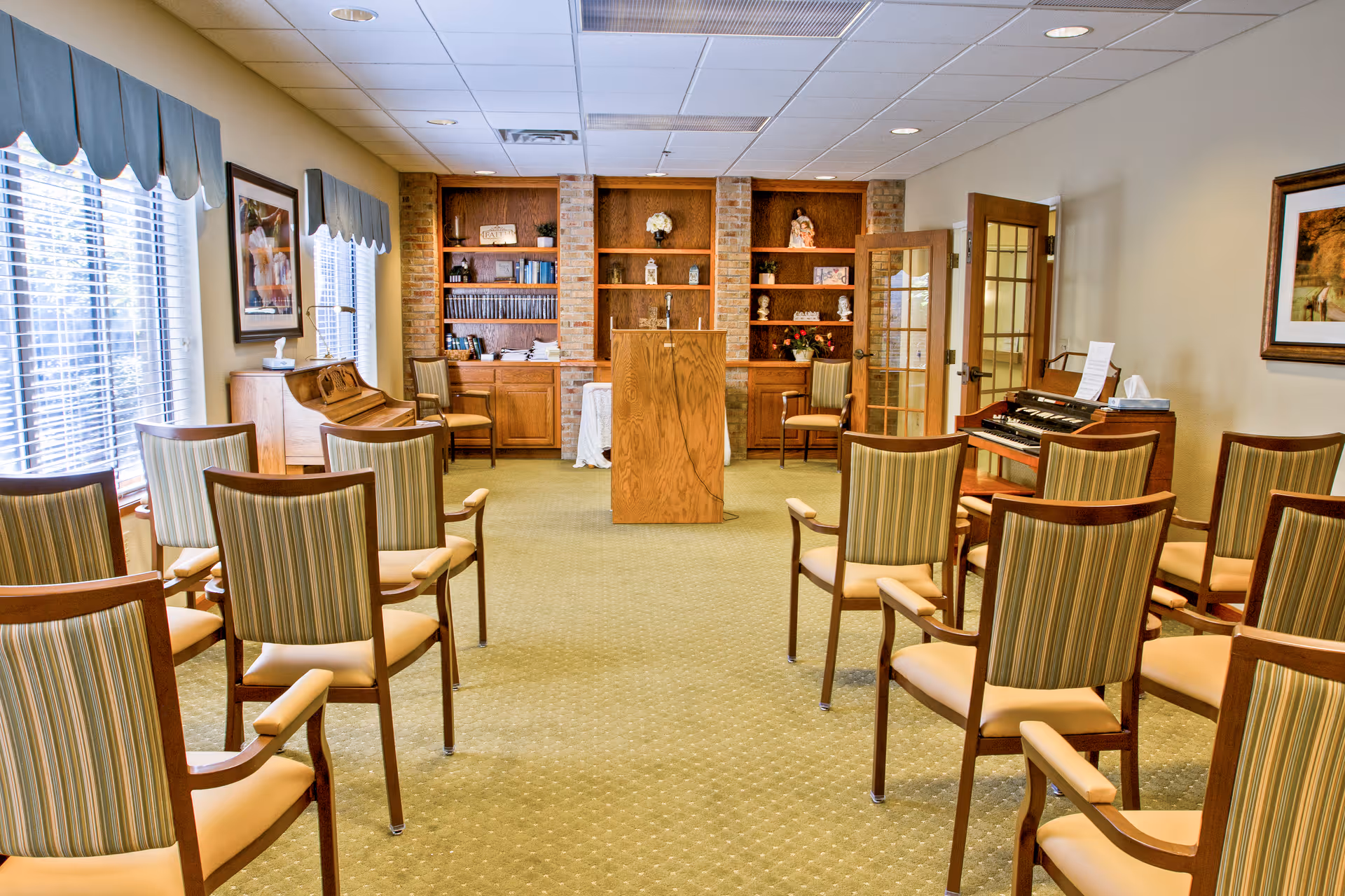A small meeting or chapel room with rows of wooden chairs with striped cushions facing a wooden podium. The room has large windows with blue valances on the left side, built-in wooden shelves with decorative items and books at the front, and a wooden organ on the right side. The walls are light-colored, and there is a framed picture hanging on the right wall.