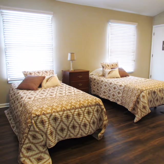 Two twin beds with matching patterned bedspreads and pillows flanking a nightstand with a lamp in a bright bedroom with two windows.
