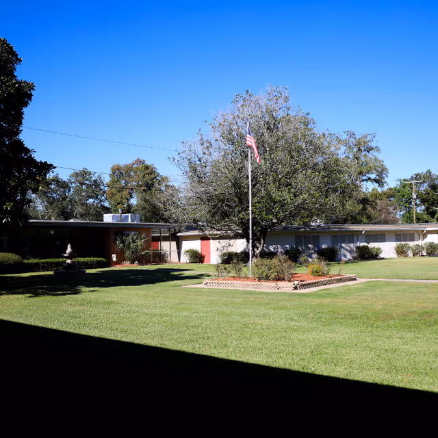 Outdoor view of a retirement home facility with a large grassy lawn, a tree with an American flag on a flagpole in the center, and a single-story building in the background under a clear blue sky.