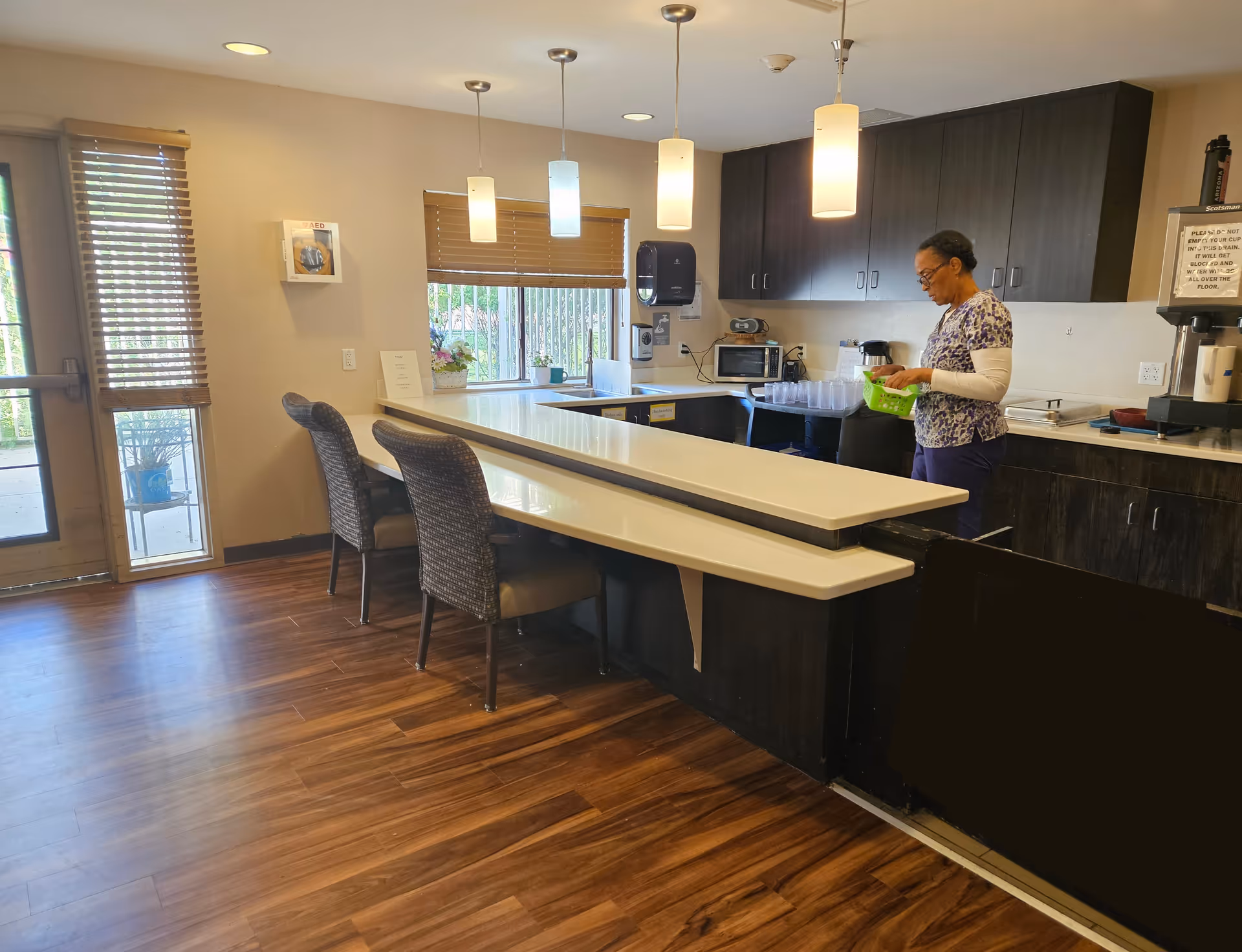 Interior view of a senior living facility kitchen area with wooden flooring, a long white countertop with two chairs, hanging pendant lights, and a woman standing behind the counter preparing items. There are dark wood cabinets, a microwave, and a coffee machine in the background.