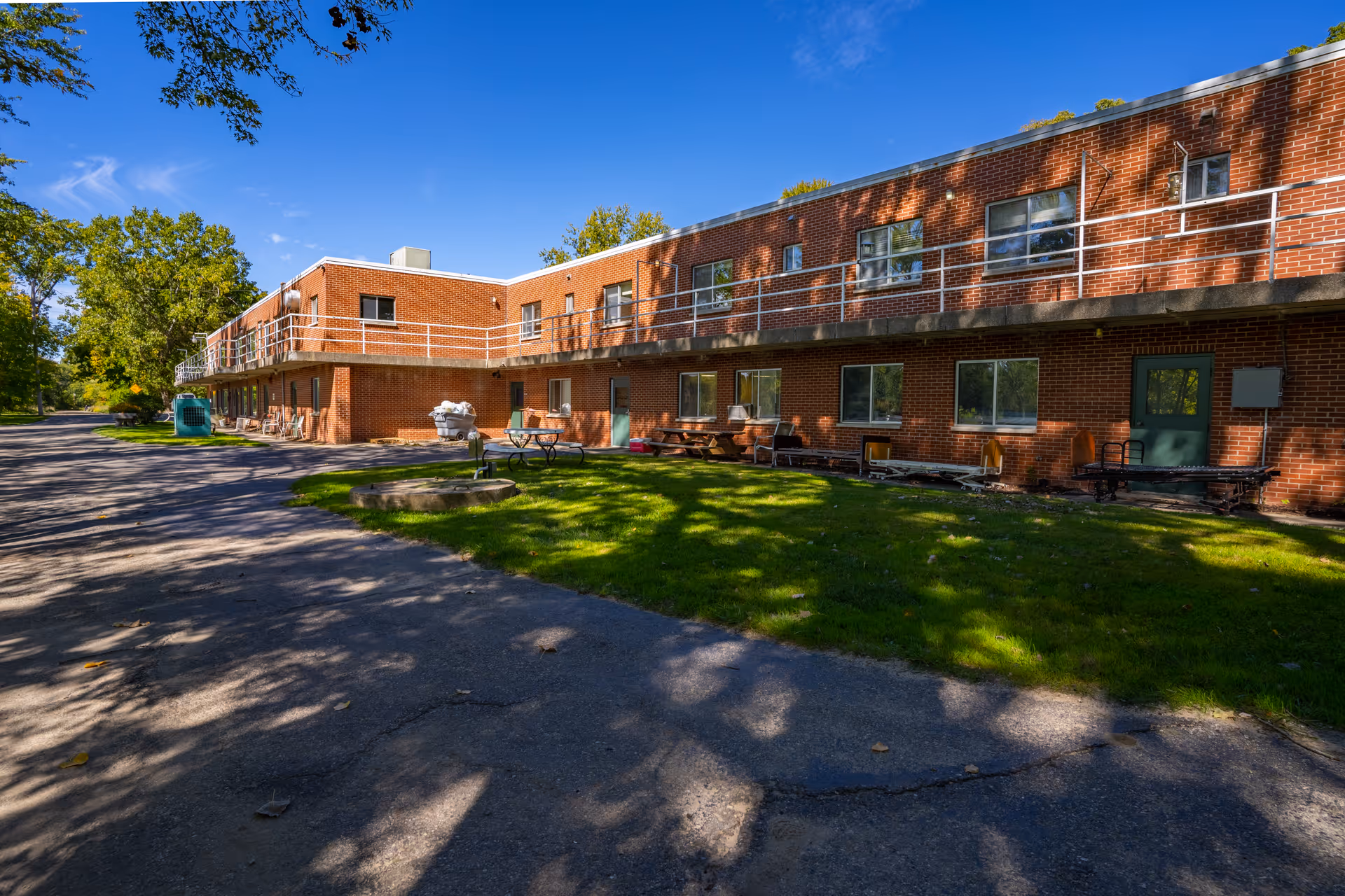 Exterior view of a two-story brick building with multiple windows and a green door, surrounded by a paved driveway and a grassy area with picnic tables and benches under a clear blue sky.