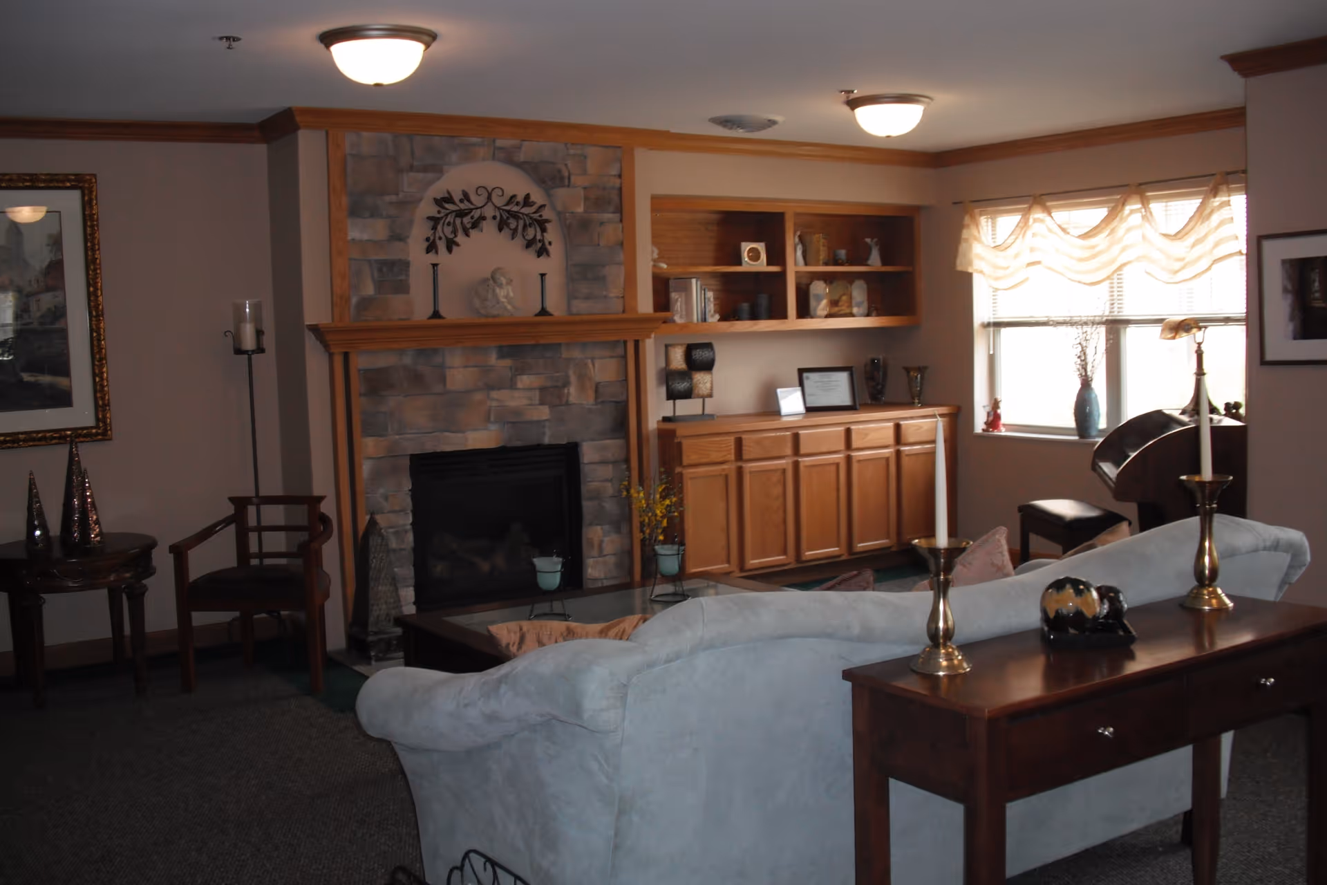A cozy living room with a light blue couch facing a stone fireplace with a wooden mantle. To the right of the fireplace is a wooden cabinet with shelves displaying decorative items. A window with sheer curtains allows natural light into the room. There is a wooden table behind the couch with two brass candle holders and a decorative item. Additional seating includes a wooden chair and a small round table with decorative cones. The room has carpeted flooring and warm lighting from ceiling fixtures.