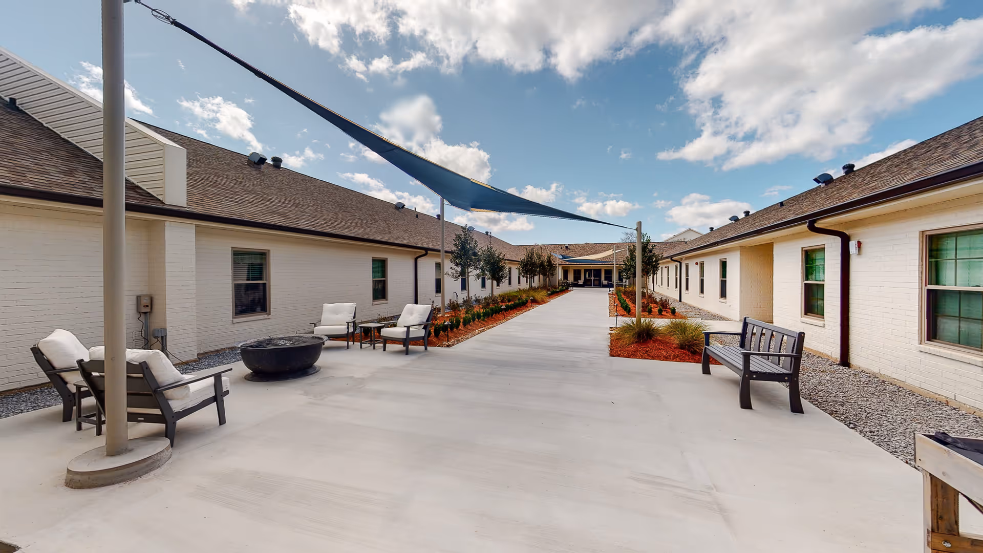 Outdoor courtyard area of a senior living facility with white brick buildings on both sides, a concrete walkway in the center, shaded seating areas with cushioned chairs and a fire pit, benches, and landscaped plants under a partly cloudy sky.