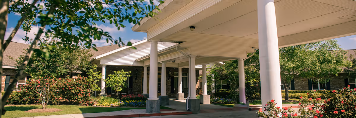 Covered entrance area of a healthcare facility with white columns, surrounded by green trees and flowering bushes under a blue sky with some clouds.