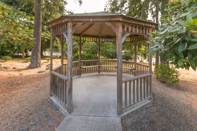 A wooden gazebo with a hexagonal shape situated outdoors surrounded by trees and shrubs, with a concrete pathway leading to its entrance.