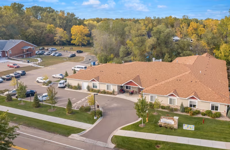 Aerial view of Suite Living Senior Care - Roseville facility showing a single-story building with a brown roof surrounded by trees with autumn foliage. There is a parking lot with several cars and a curved driveway leading to the entrance. A sign for Suite Living Senior Care is visible on the lawn near the sidewalk.