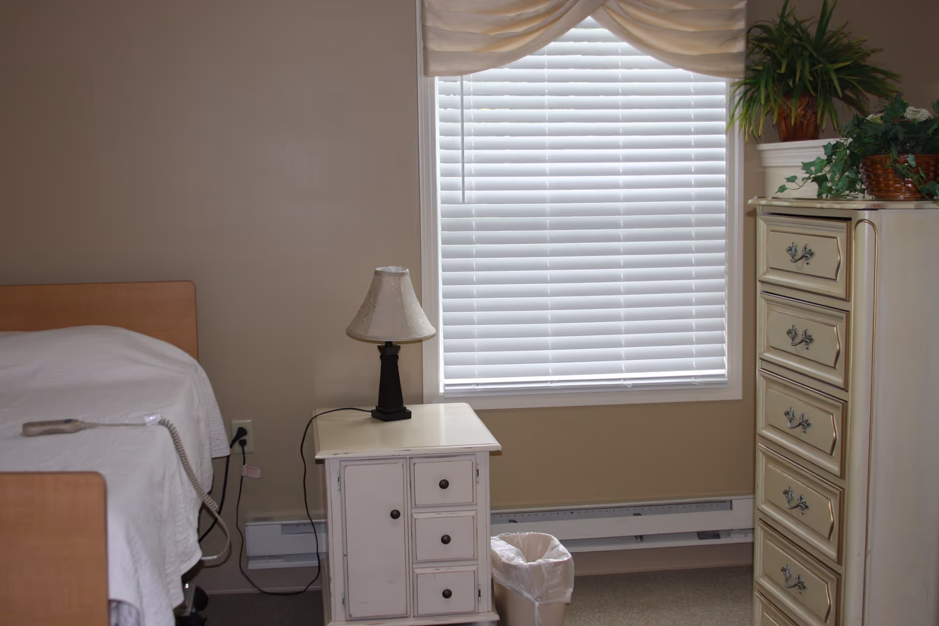 A simple bedroom with a bed covered in a white quilt, a white nightstand with a lamp, a window with white blinds and a valance, a trash can, and a cream-colored chest of drawers with plants on top.