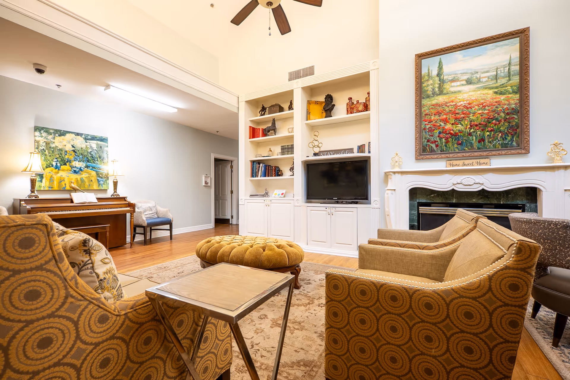 A cozy senior living room with patterned armchairs, a round tufted ottoman, a fireplace with a colorful landscape painting above it, built-in shelves with books and decorative items, a flat-screen TV, a piano with a painting above it, and a ceiling fan.