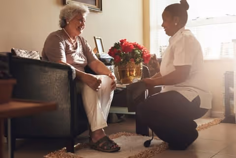 An elderly woman sitting in a chair smiling while a caregiver kneels in front of her, helping to put on or adjust her shoe in a warmly lit room with a window and a vase of red flowers on a table.