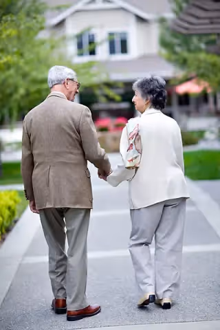 An elderly couple holding hands and walking on a paved pathway in a residential or senior living community with greenery and buildings in the background.