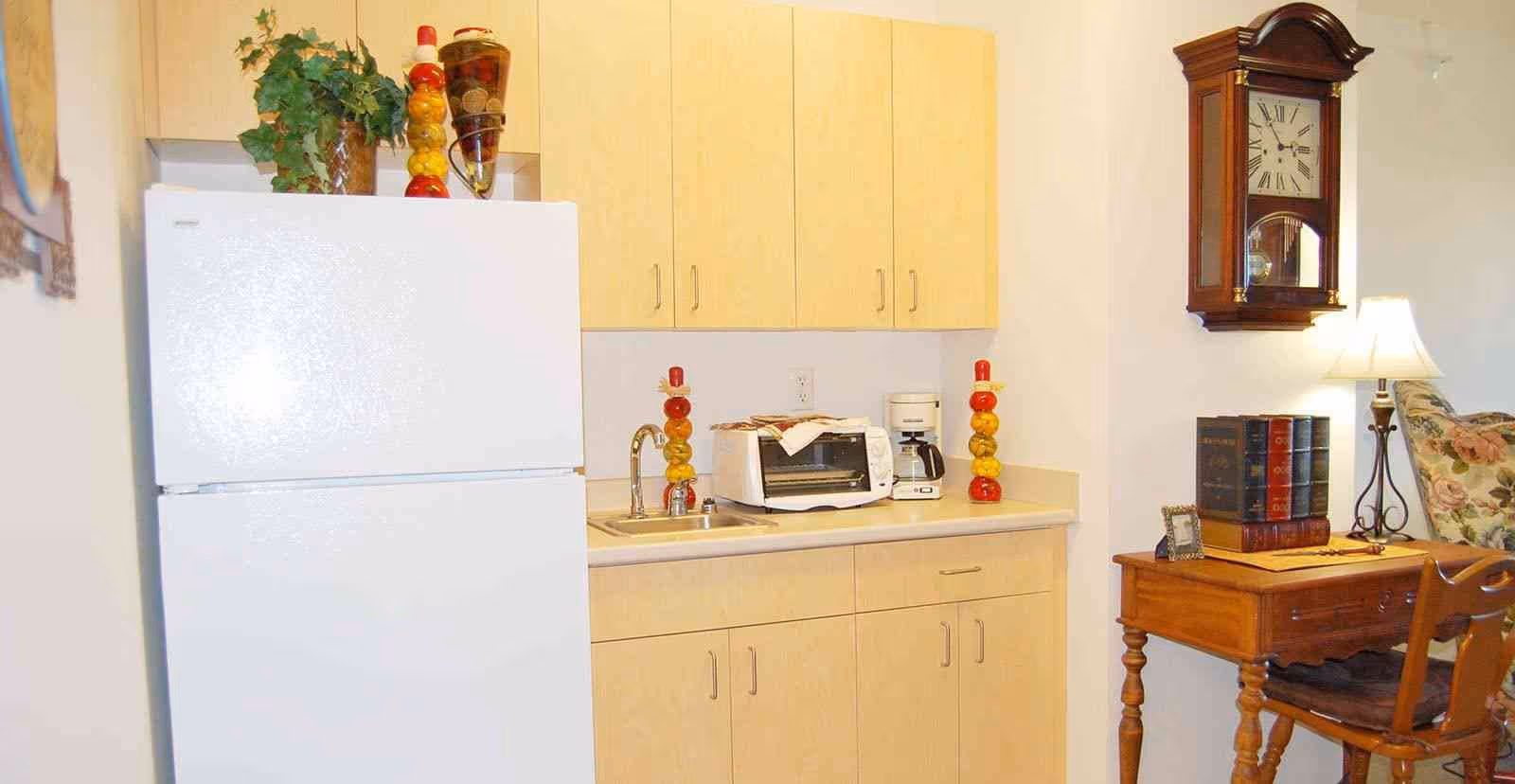 Small kitchenette with a white refrigerator, light wood cabinets, sink, toaster and coffee maker beside a wooden side table and wall clock.