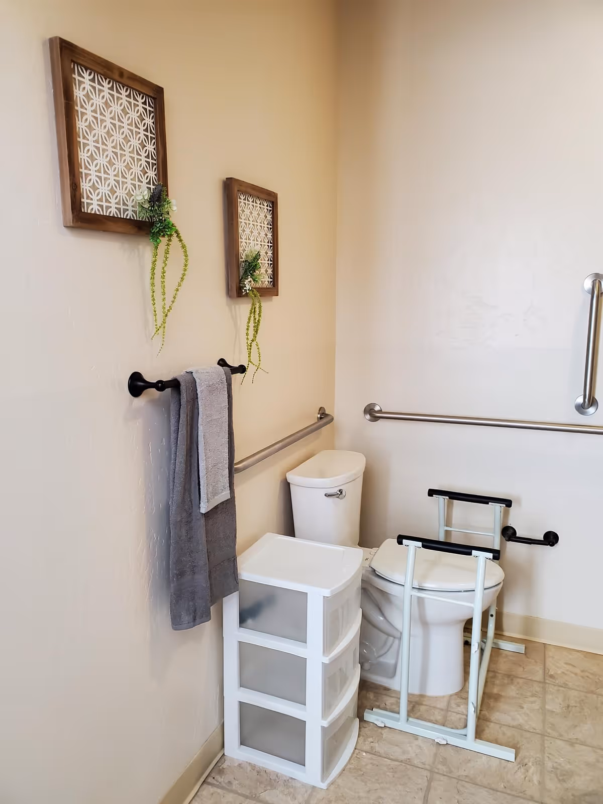 A bathroom corner featuring a white toilet with safety grab bars on the walls and a white support frame around the toilet. Next to the toilet is a white plastic storage drawer unit. Two framed decorative wall hangings with green plants are mounted above a black towel rack holding two gray towels. The floor is tiled in a light beige color.