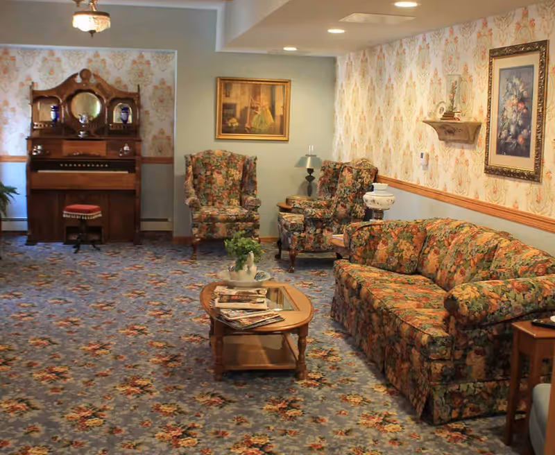 A cozy living room in a senior living facility featuring floral patterned carpet and matching floral upholstered sofa and armchairs. There is a wooden coffee table with magazines and a decorative plant, a vintage wooden piano with a red cushioned stool, and framed artwork on the walls with floral wallpaper.