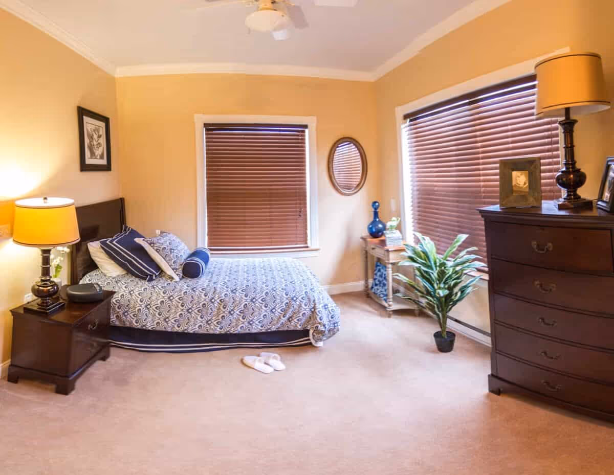 A cozy bedroom with beige walls and carpet, featuring a bed with patterned blue and white bedding, a nightstand with a lamp and a small plant, a wooden dresser with a lamp and framed pictures, two windows with closed brown blinds, a small table with decorative items, a round wall mirror, and a potted plant on the floor.