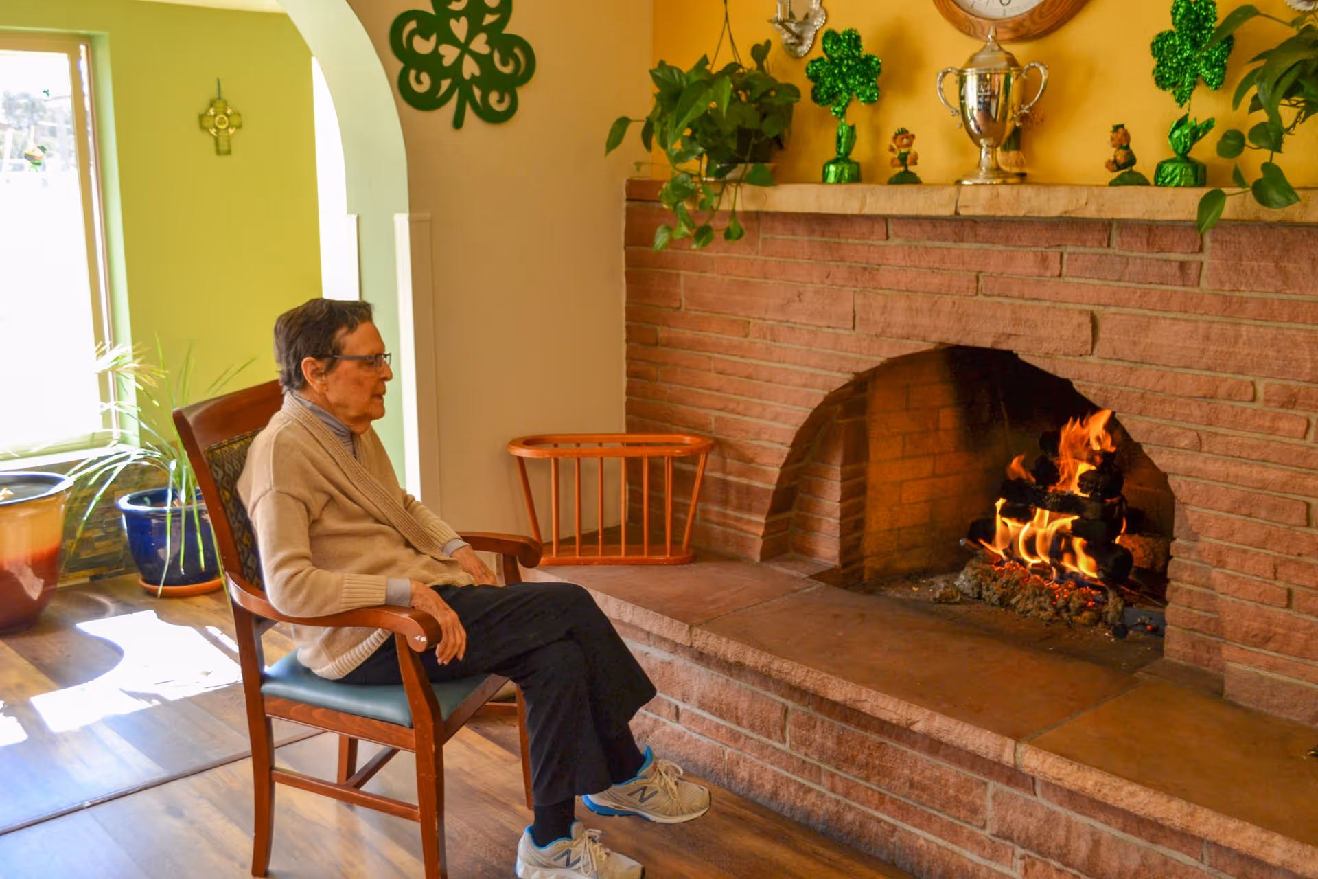 An elderly person sitting in a wooden chair facing a lit brick fireplace in a cozy room decorated with green shamrock ornaments and potted plants.