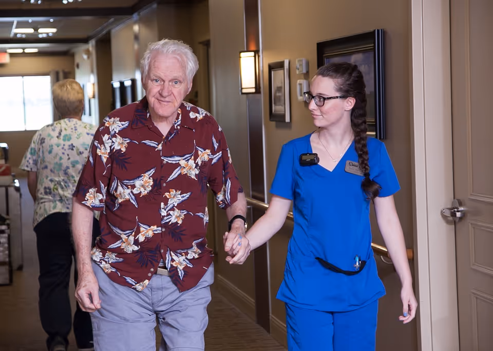 An elderly man wearing a floral shirt is walking down a hallway holding hands with a female caregiver dressed in blue scrubs. Another person is walking ahead in the hallway. The setting appears to be inside a senior living facility.