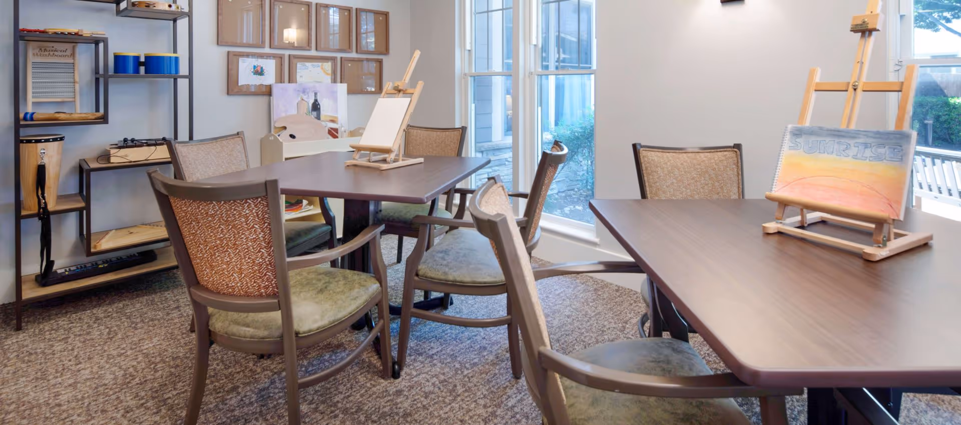 A bright activity room with two wooden tables surrounded by chairs. Each table has a small wooden easel, one holding a blank canvas and the other displaying a painting with the word 'SUNRISE'. Shelves on the left wall hold musical instruments and art supplies. Large windows let in natural light and show greenery outside.
