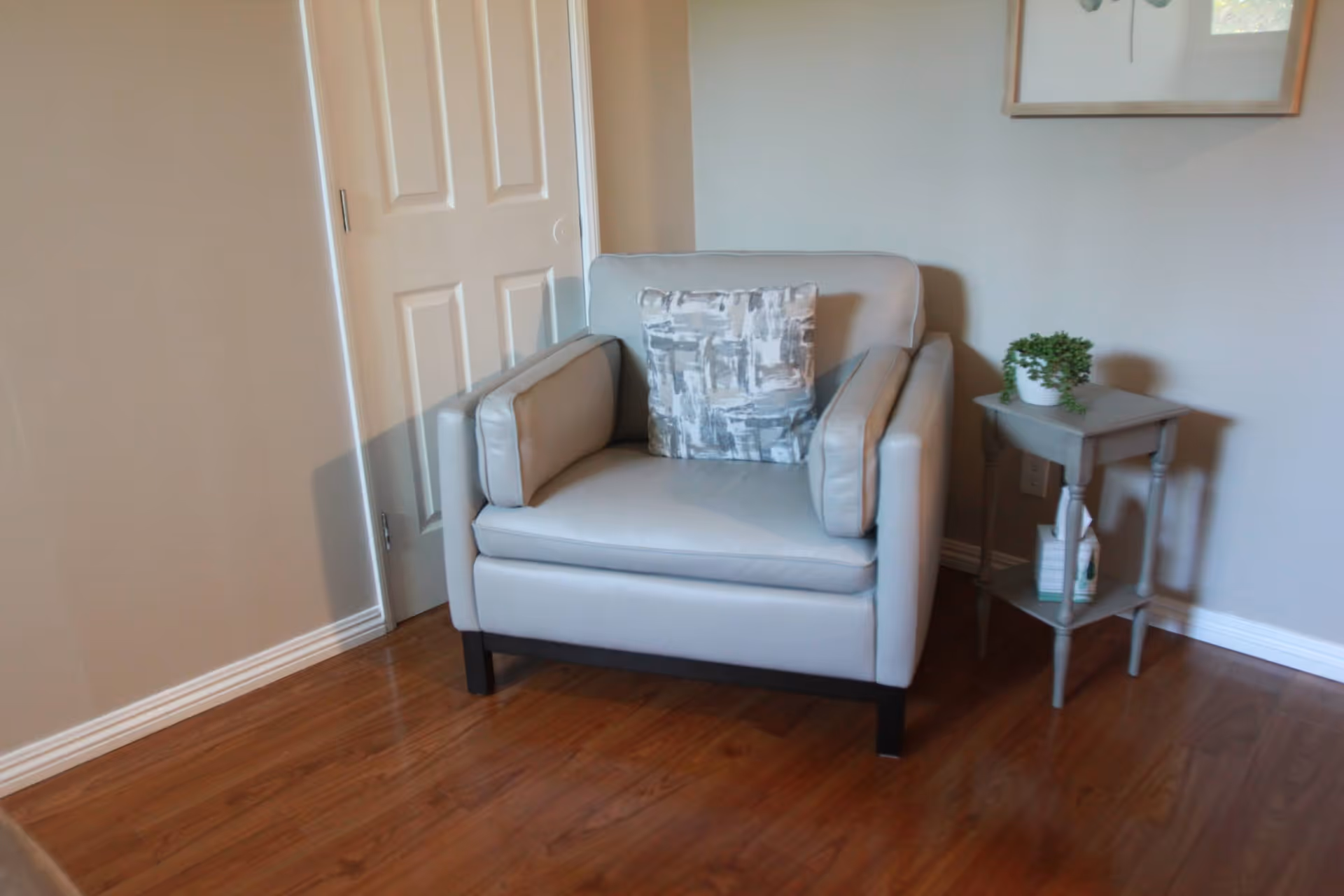 A light gray armchair with a patterned cushion is placed on a wooden floor next to a small gray side table holding a potted plant and some books, with a closed white door and a framed picture on the wall in the background.