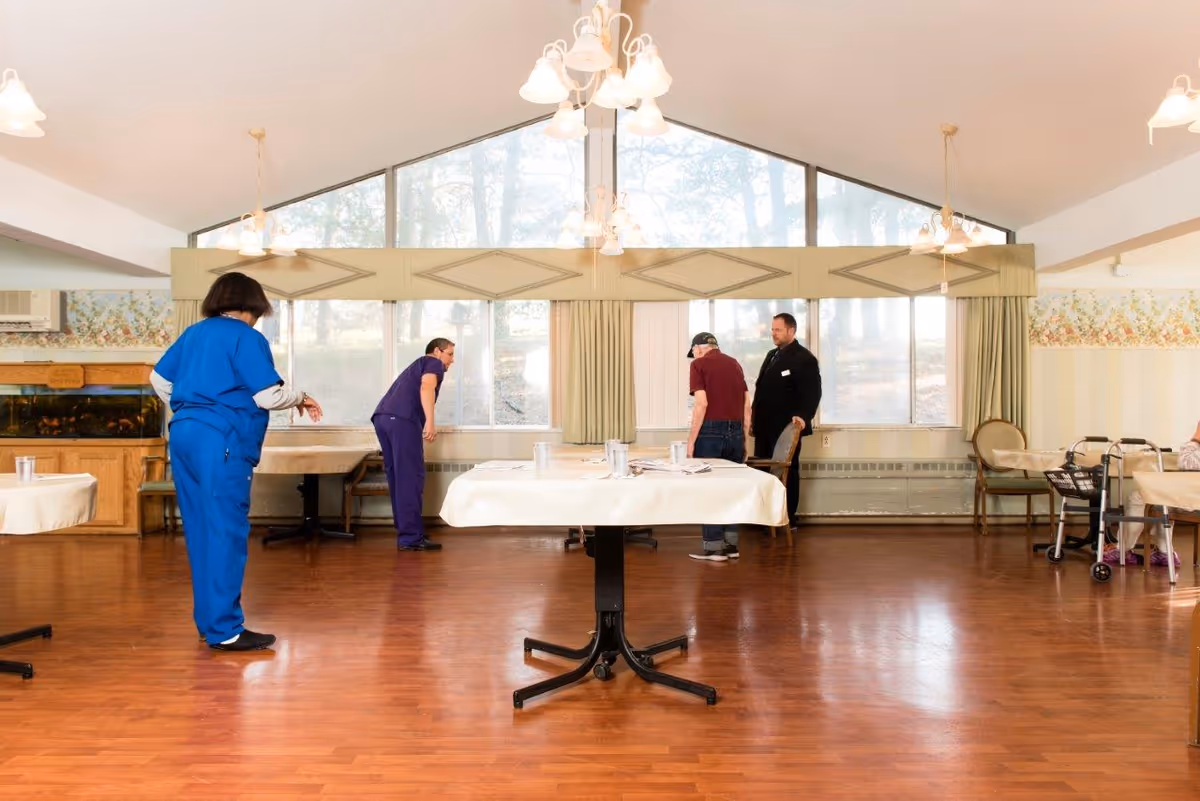 A spacious dining room with wooden floors and large windows letting in natural light. There are several tables covered with white tablecloths, and a few people are present, including two staff members in blue uniforms and two elderly individuals, one using a walker. The room has floral wallpaper and multiple hanging light fixtures.