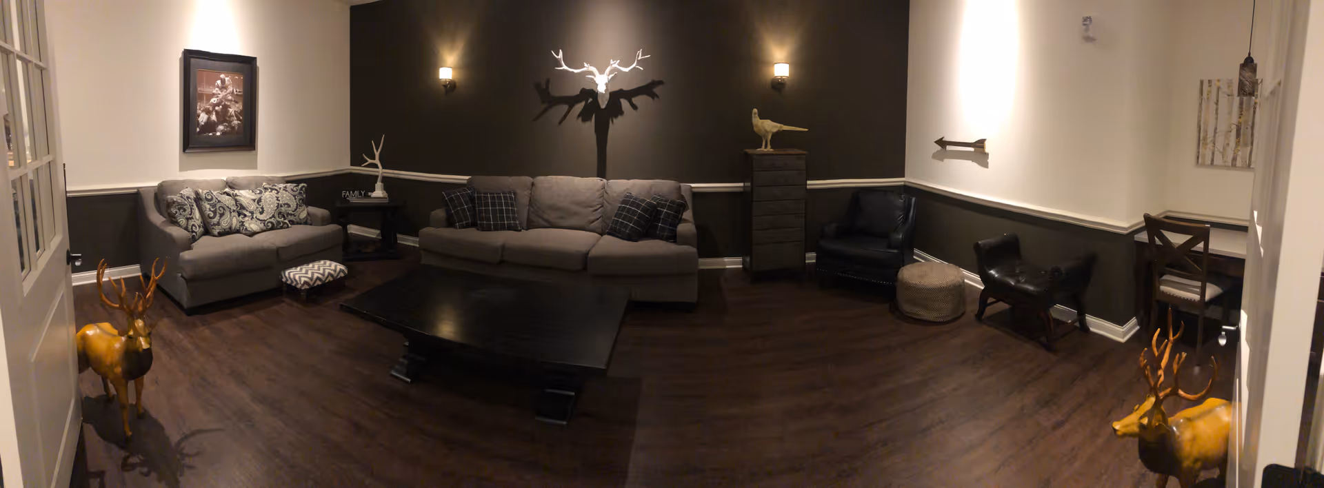 A cozy living room with dark wood flooring and two-tone walls painted white and dark brown. The room features a gray sofa with patterned cushions, a matching loveseat, a black coffee table, two black armchairs, a small side table with decorative items, and two decorative deer statues near the entrance. Wall sconces provide soft lighting, and there is a framed picture and a wall-mounted deer head decoration.