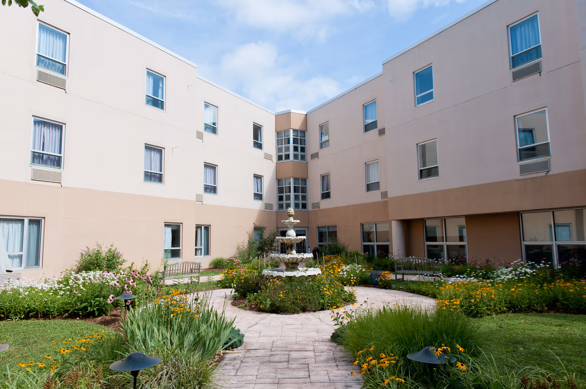 A landscaped courtyard with a multi-tiered fountain, flowering gardens and benches surrounded by a three-story senior living building.