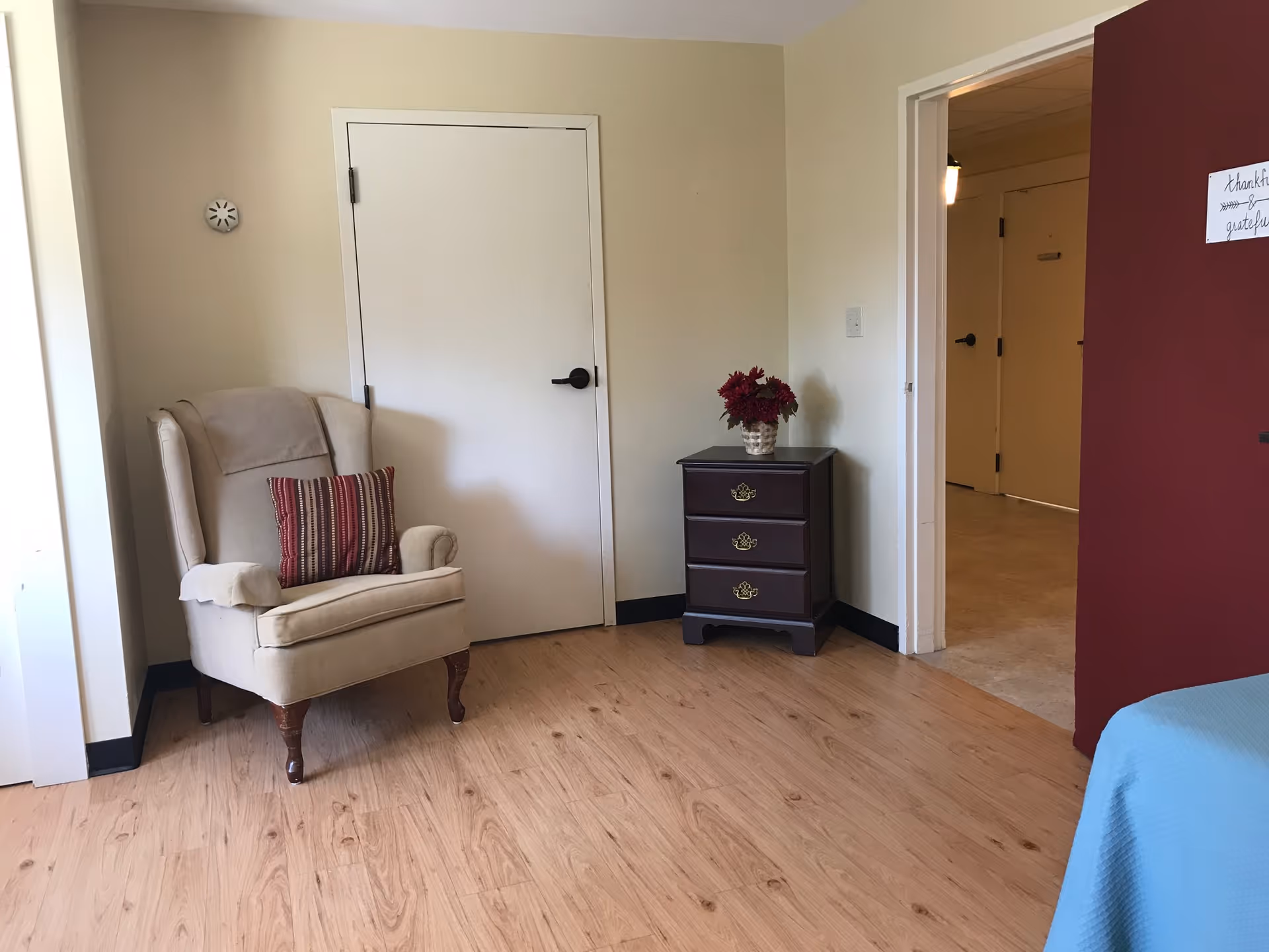 A cozy corner of a room featuring a beige upholstered armchair with a striped pillow, a small dark wooden three-drawer nightstand with a vase of red flowers on top, light wood flooring, a closed white door, and a partial view of a bed with a blue blanket. The walls are painted in light beige and dark red, with a small sign on the red wall that reads 'thankful grateful'.