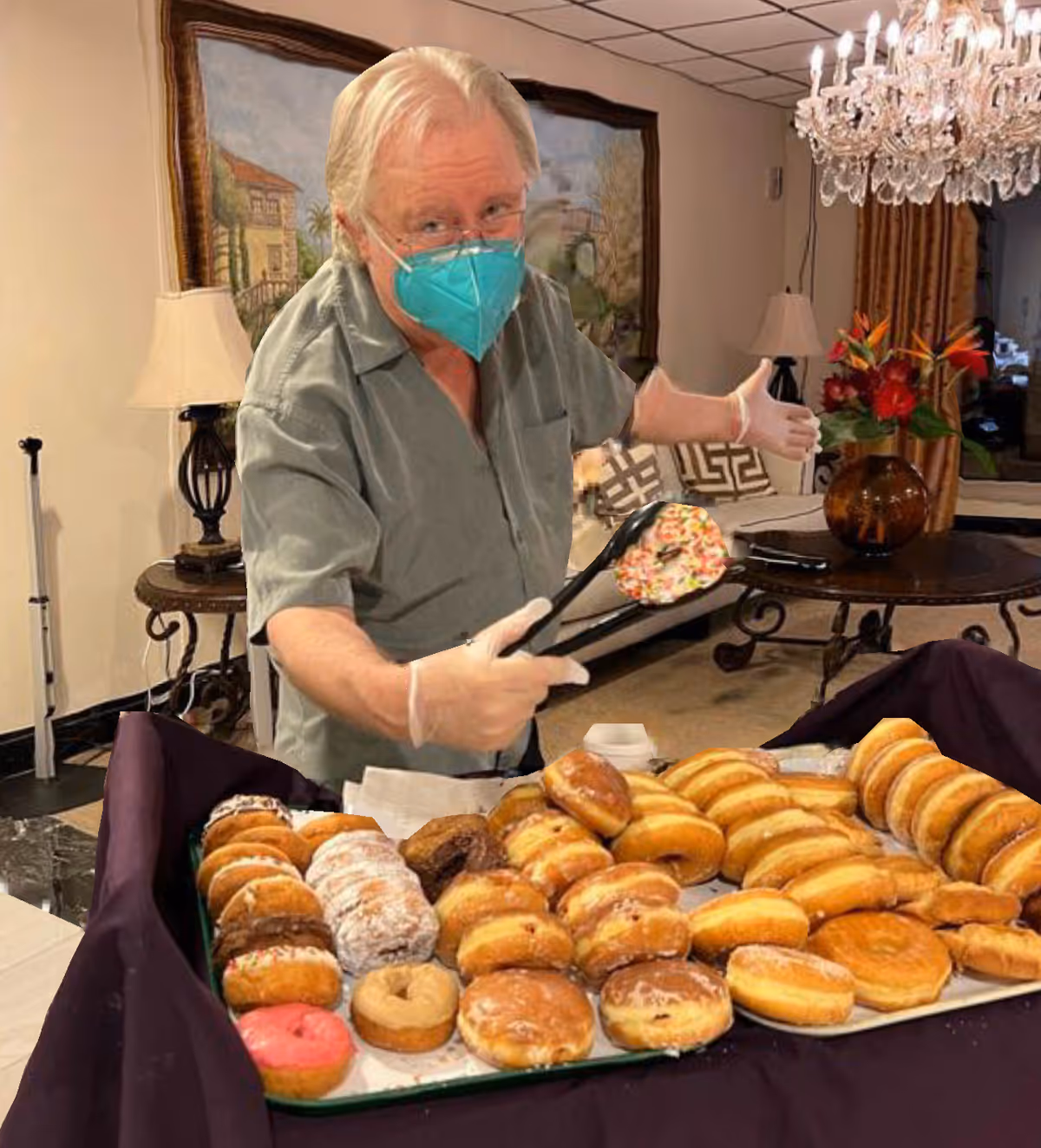 An elderly man wearing a teal face mask and gloves is holding tongs with a sprinkled donut in a room with a chandelier, a table with a vase of flowers, and a painting on the wall. In front of him is a tray filled with various types of donuts.
