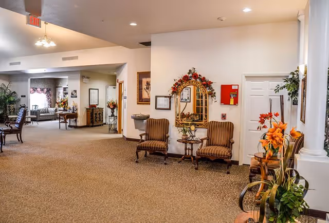 Spacious furnished senior living common area with upholstered chairs, a decorative mirror, and floral arrangements.