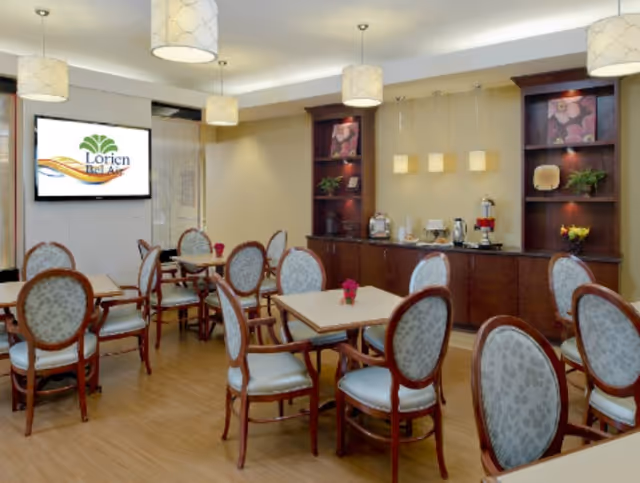 Bright dining area with multiple wooden tables and upholstered chairs, a buffet counter along the back wall, and a wall-mounted screen showing the Lorien Bel Air logo.