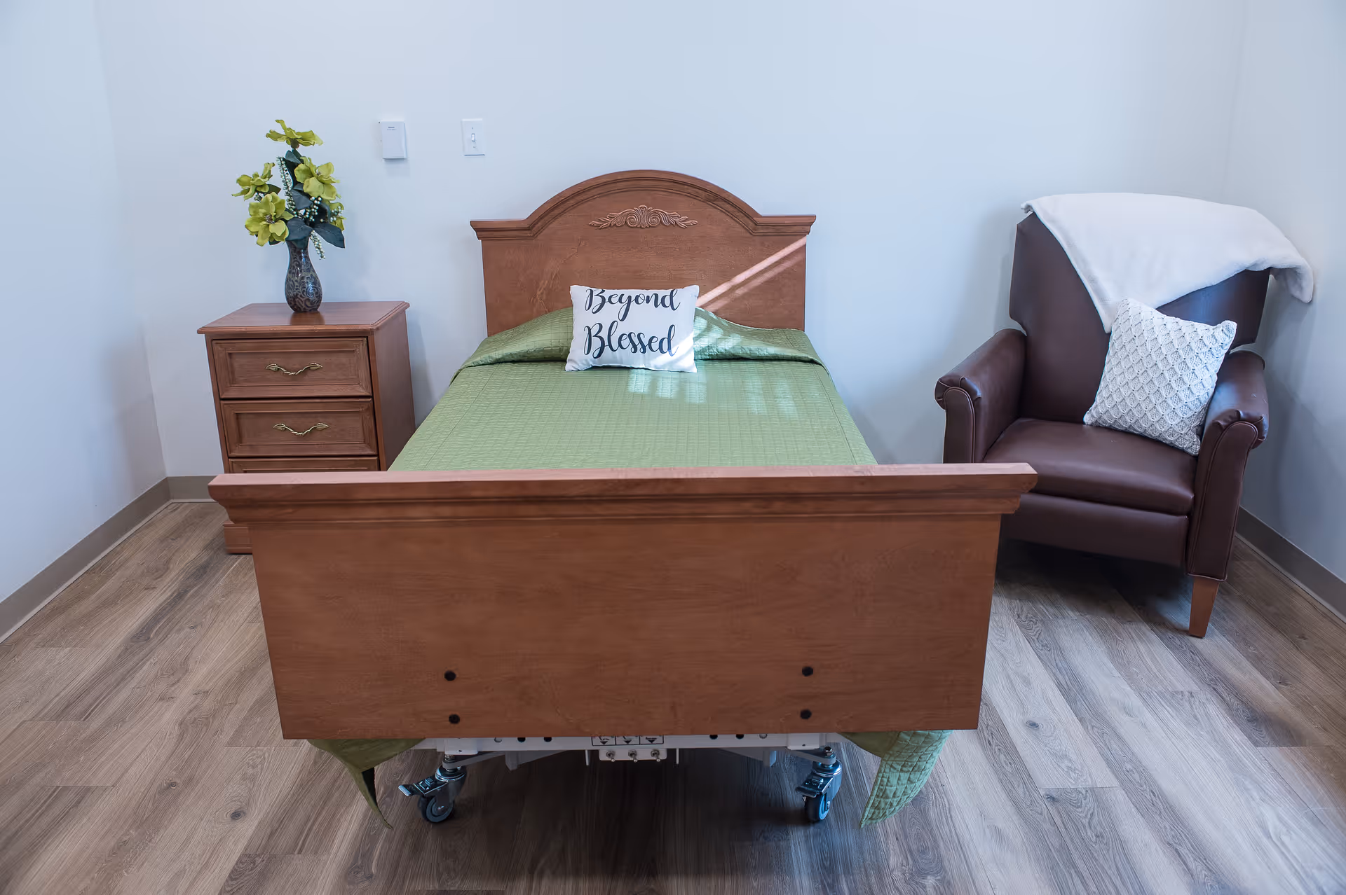 A neatly made single bed with a wooden headboard and footboard, covered with a green quilt and a decorative pillow that says 'Beyond Blessed'. To the left of the bed is a wooden nightstand with two drawers and a vase with yellow flowers. To the right of the bed is a brown leather armchair with a white knitted pillow and a white throw draped over the back. The room has light-colored walls and wood flooring.