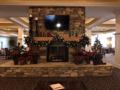 Stone fireplace in a communal lounge decorated for the holidays with a TV above, garlands, poinsettias, and dining tables visible in the background.
