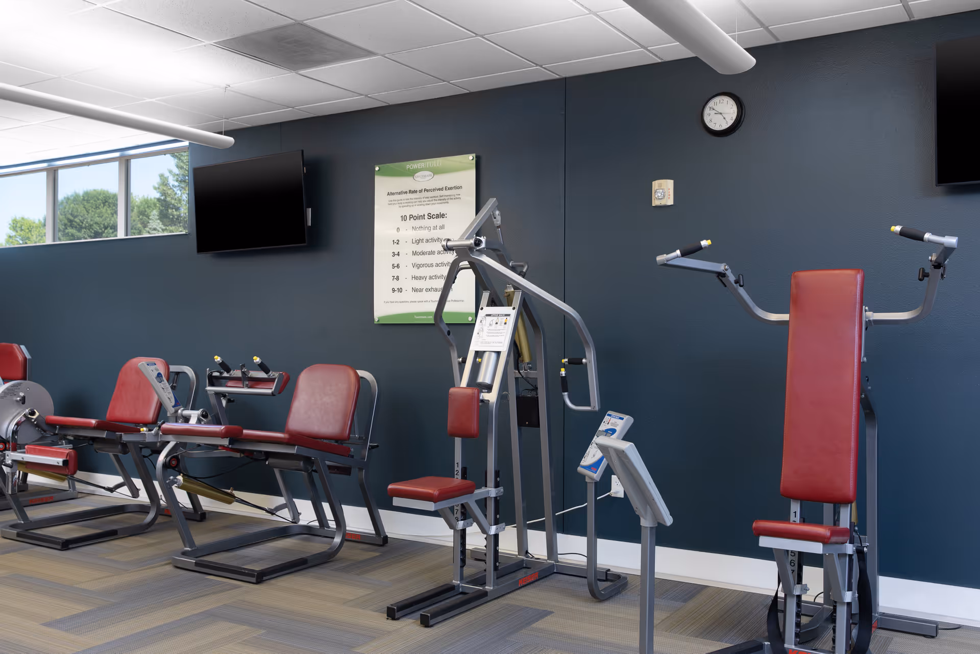 A fitness room with several exercise machines featuring red padded seats and backrests, set against a dark blue wall. There are two flat-screen TVs mounted on the wall, a clock, and a poster displaying a 10 point scale for perceived exertion. The room has a window letting in natural light and a patterned carpeted floor.