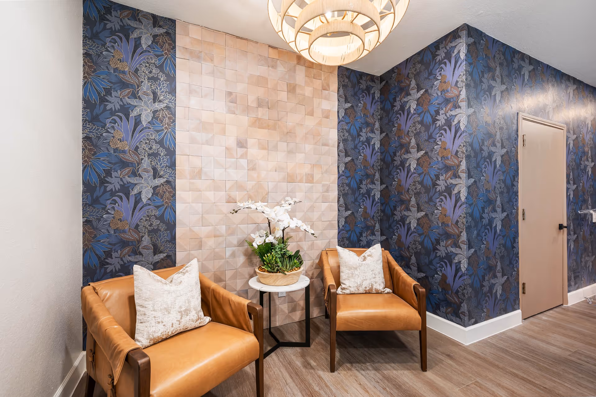 A cozy seating area with two tan leather armchairs, each with a light-colored pillow, positioned on either side of a small round table holding a white orchid plant. The walls feature a combination of beige textured tiles and dark blue floral wallpaper. A modern circular chandelier hangs from the ceiling, and a closed beige door is visible on the right side.