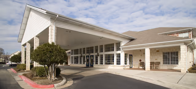 Exterior view of the entrance to a senior living facility with a covered drop-off area supported by stone columns, large windows, and double doors under a partly cloudy sky.