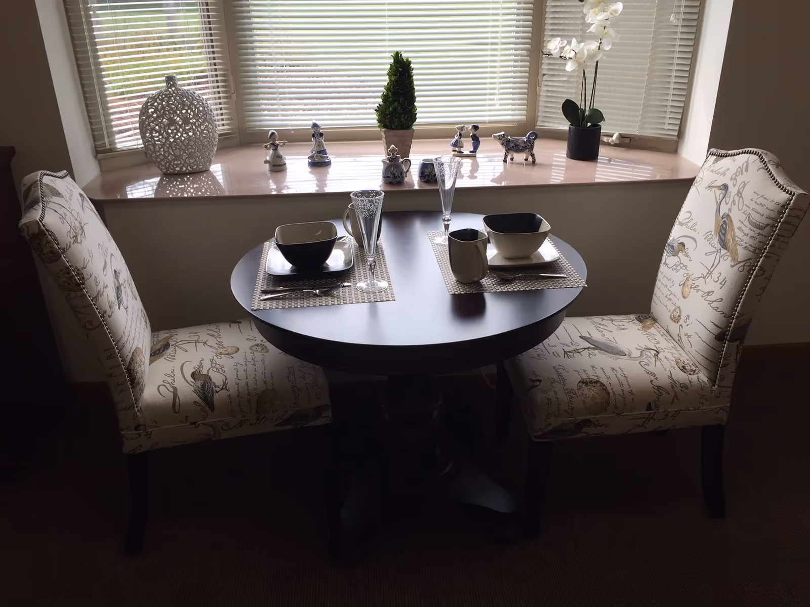 A small round dining table set for two with matching patterned chairs. The table has two place settings with bowls, plates, mugs, and glasses. Behind the table is a window with blinds and a windowsill decorated with small figurines, a potted plant, and a white decorative vase.