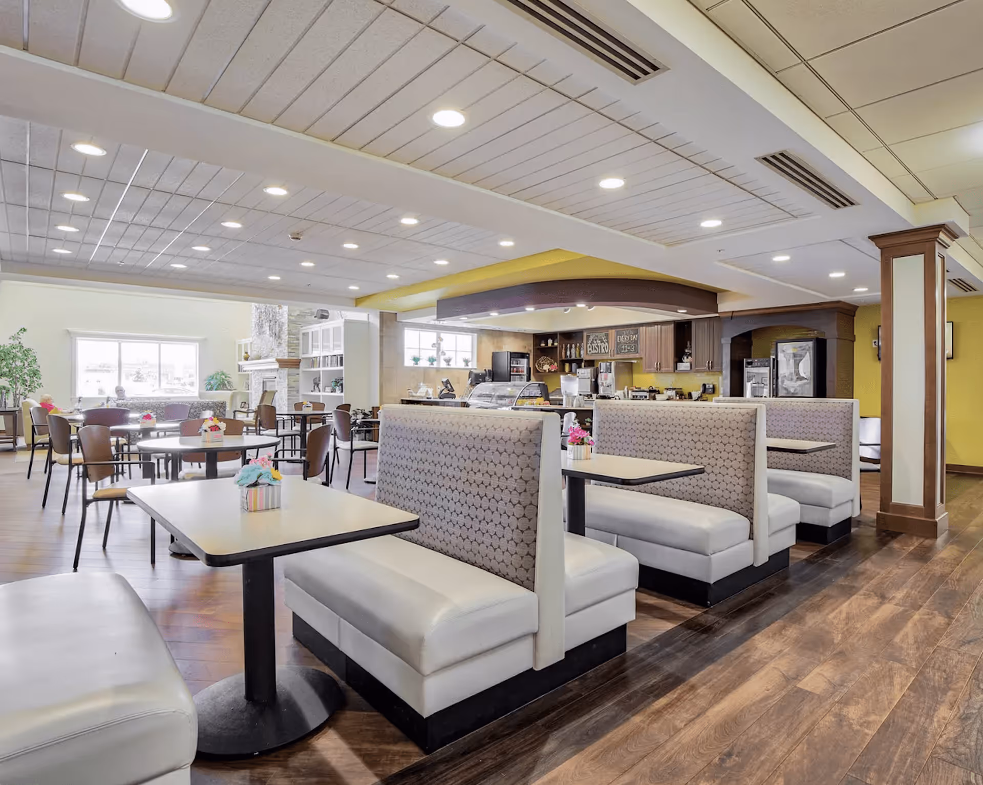 Interior view of a dining area in a senior living facility with booth seating and tables. The space features wooden flooring, a counter area with a coffee machine and display case, and large windows letting in natural light. There are also round tables with chairs and a fireplace in the background.
