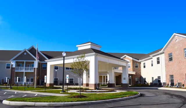 Exterior view of Danbury Huber Heights senior living facility showing a large building with beige and brick walls, multiple windows, and a covered entrance with a driveway. The sky is clear and blue, and there are small trees and a flagpole near the entrance.