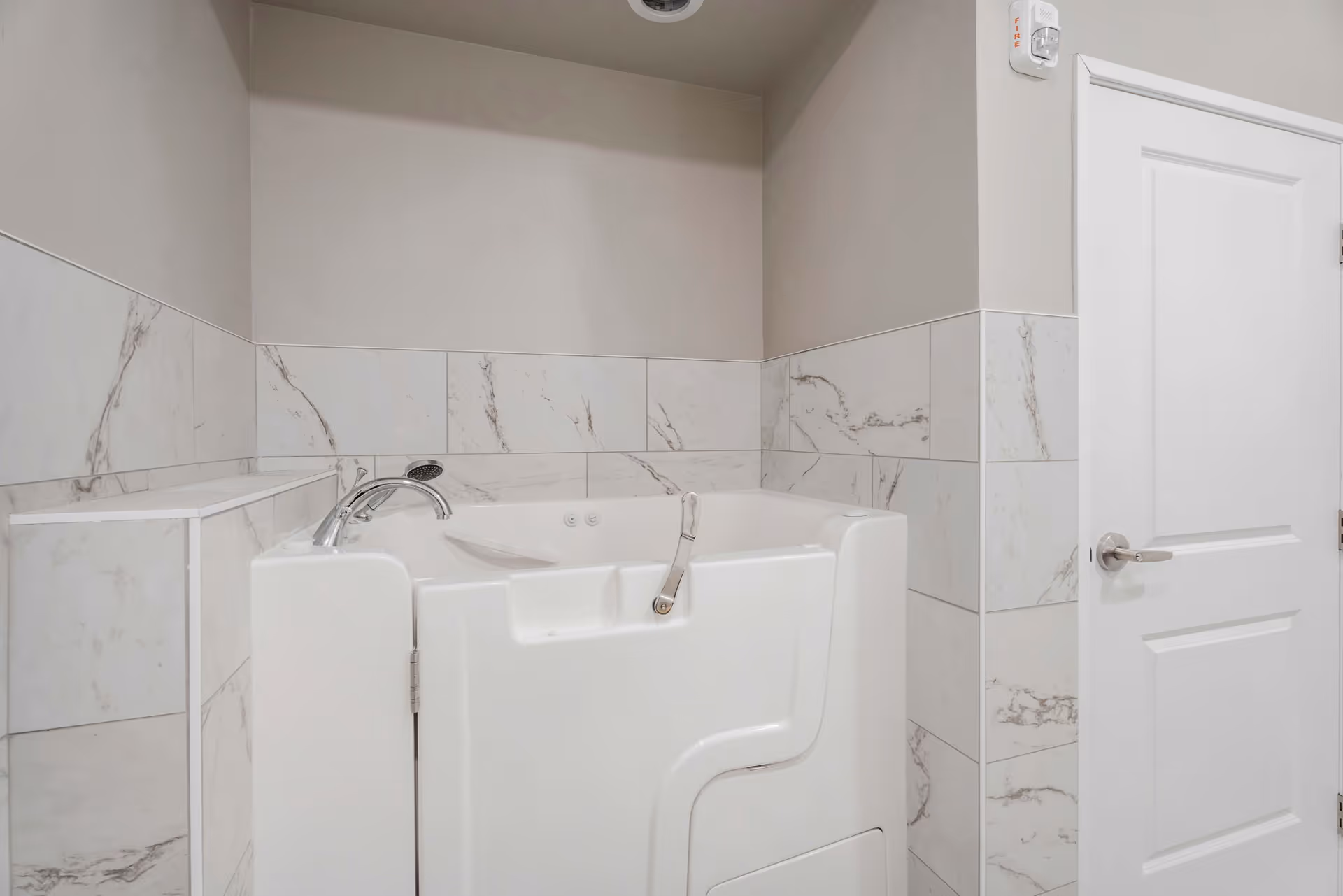 A white walk-in bathtub with chrome fixtures set against marble-patterned tiled walls next to a closed white door.