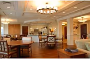 A spacious and well-lit common area in a senior living facility featuring multiple wooden tables and chairs, a kitchen area with appliances and cabinetry in the background, and comfortable seating including a sofa. The room has warm lighting with ceiling fixtures and a wooden floor.