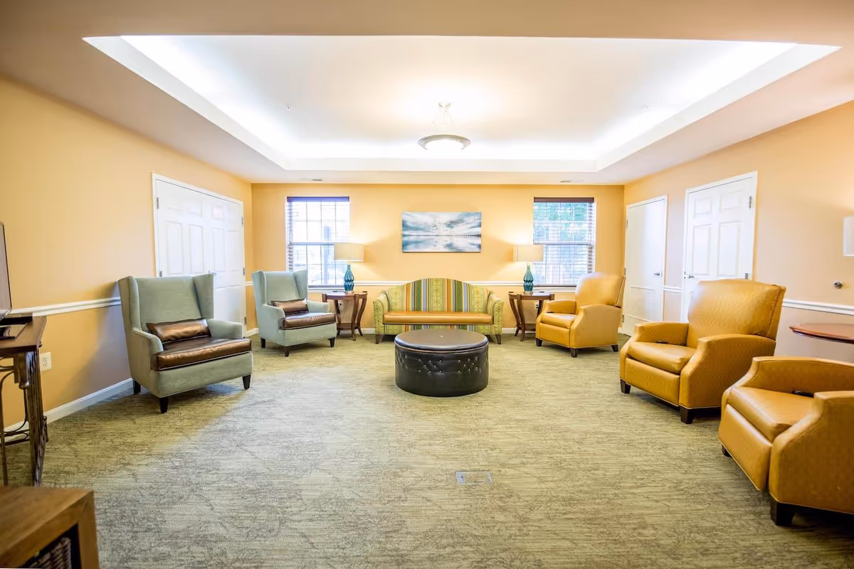 A spacious living room with beige walls and carpeted floor, featuring a green striped sofa centered between two windows with blinds. On either side of the sofa are wooden side tables with blue glass lamps. There are two light blue armchairs with brown cushions on the left and three mustard yellow armchairs on the right. A round black ottoman is placed in the center of the room. The ceiling has recessed lighting and a central hanging light fixture.