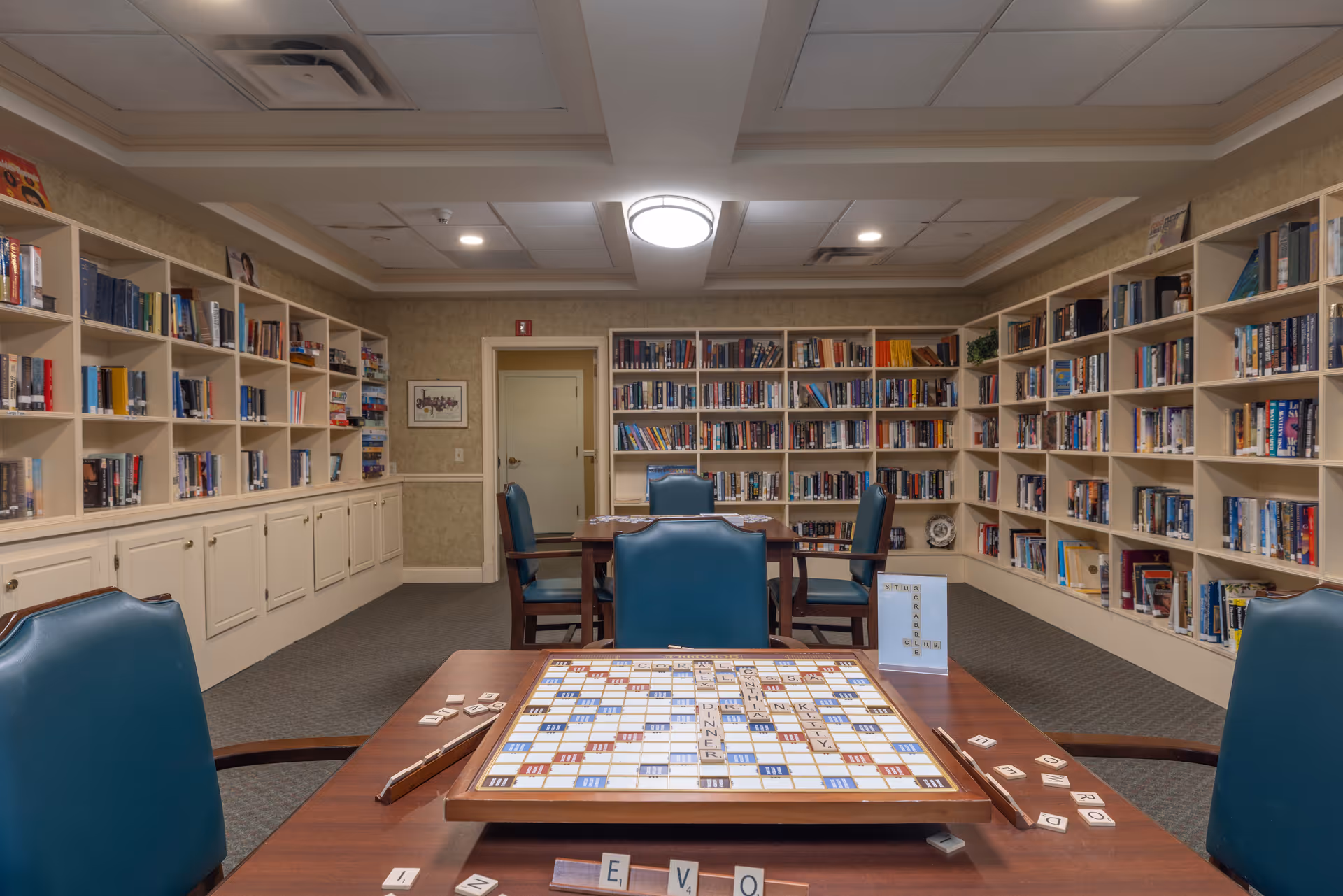 A cozy library room with bookshelves lining the walls filled with books. In the center, there are tables and chairs with a Scrabble board game set up on one table. The room has a carpeted floor and a ceiling light fixture.