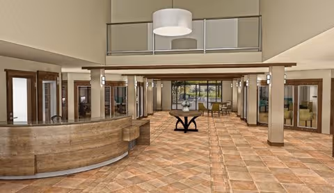 Spacious senior living facility lobby with a curved wooden reception desk on the left, a central table with a flower arrangement, tiled flooring, and multiple seating areas visible through glass doors. The ceiling is high with a modern light fixture hanging above.