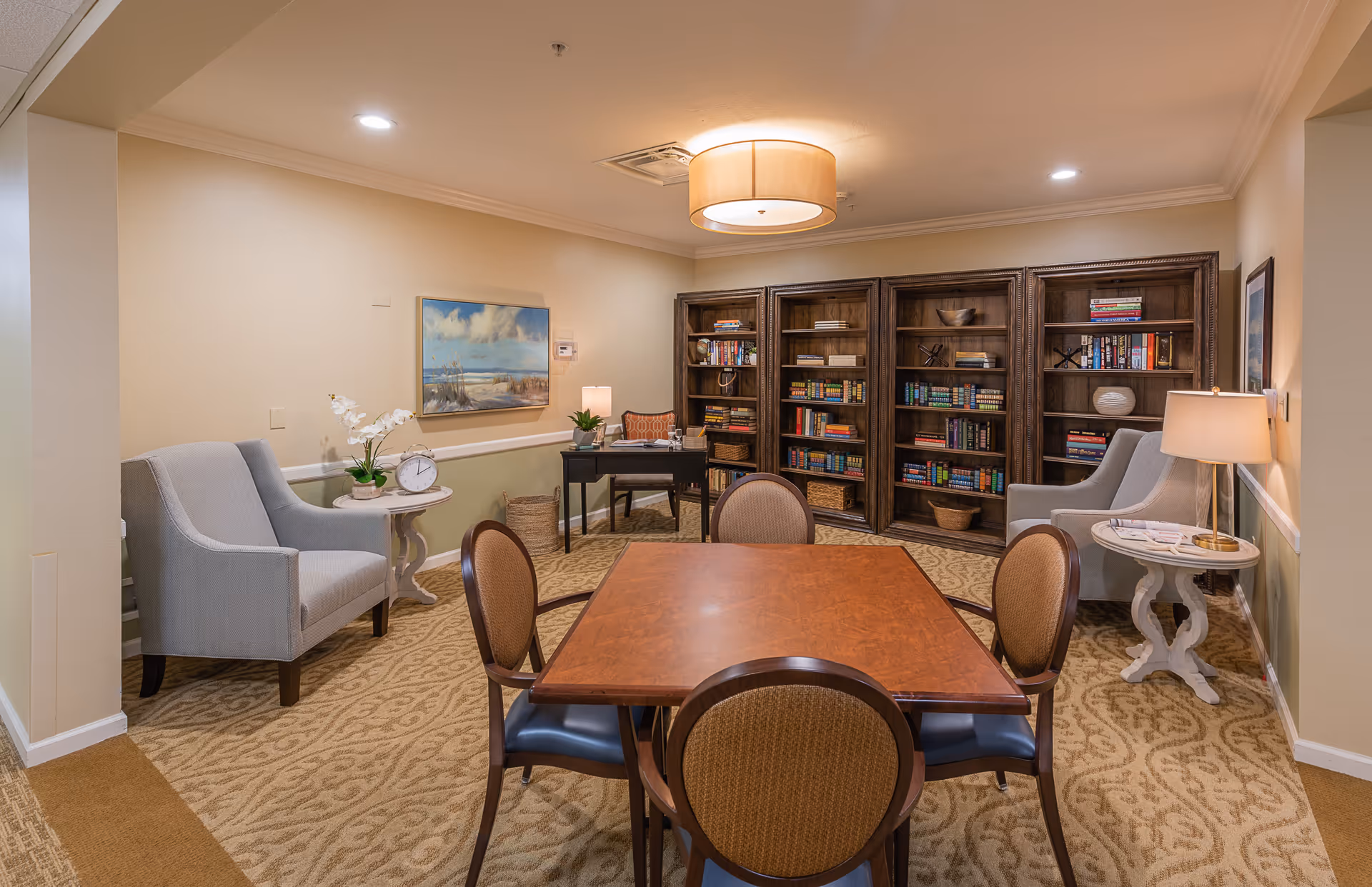 A cozy interior room with a square wooden table surrounded by four chairs in the center. Behind the table, there are three large wooden bookshelves filled with books and decorative items. On either side of the bookshelves, there are two gray armchairs next to small round side tables with lamps and decorative plants. The walls are painted beige with a framed landscape painting on the left wall. The room is softly lit by ceiling lights and a central hanging lamp.