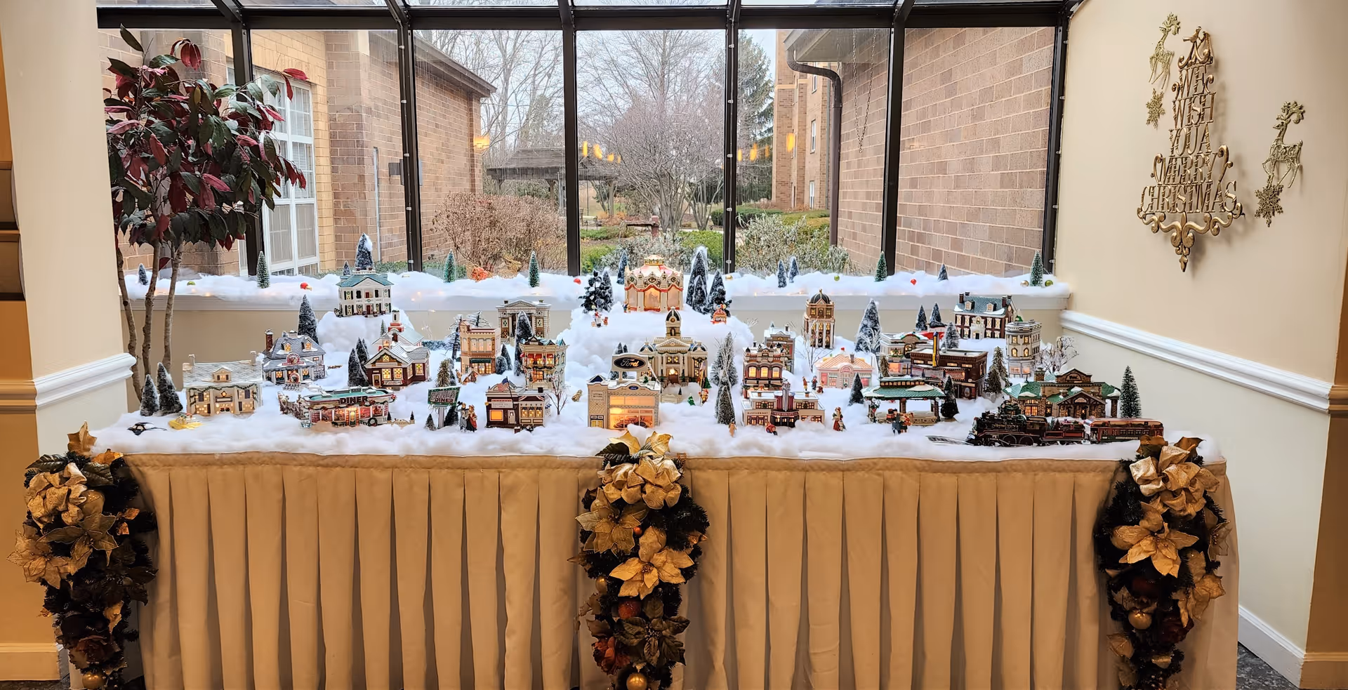 A festive miniature Christmas village display set on a table covered with a beige skirt. The village includes small buildings, trees, and a train, all surrounded by artificial snow. The table is decorated with gold and brown poinsettia garlands on the corners. Behind the display is a large window showing an outdoor garden area. On the right wall, there is a gold decorative sign that reads 'A Merry Wish For A Merry Christmas' with reindeer figures.