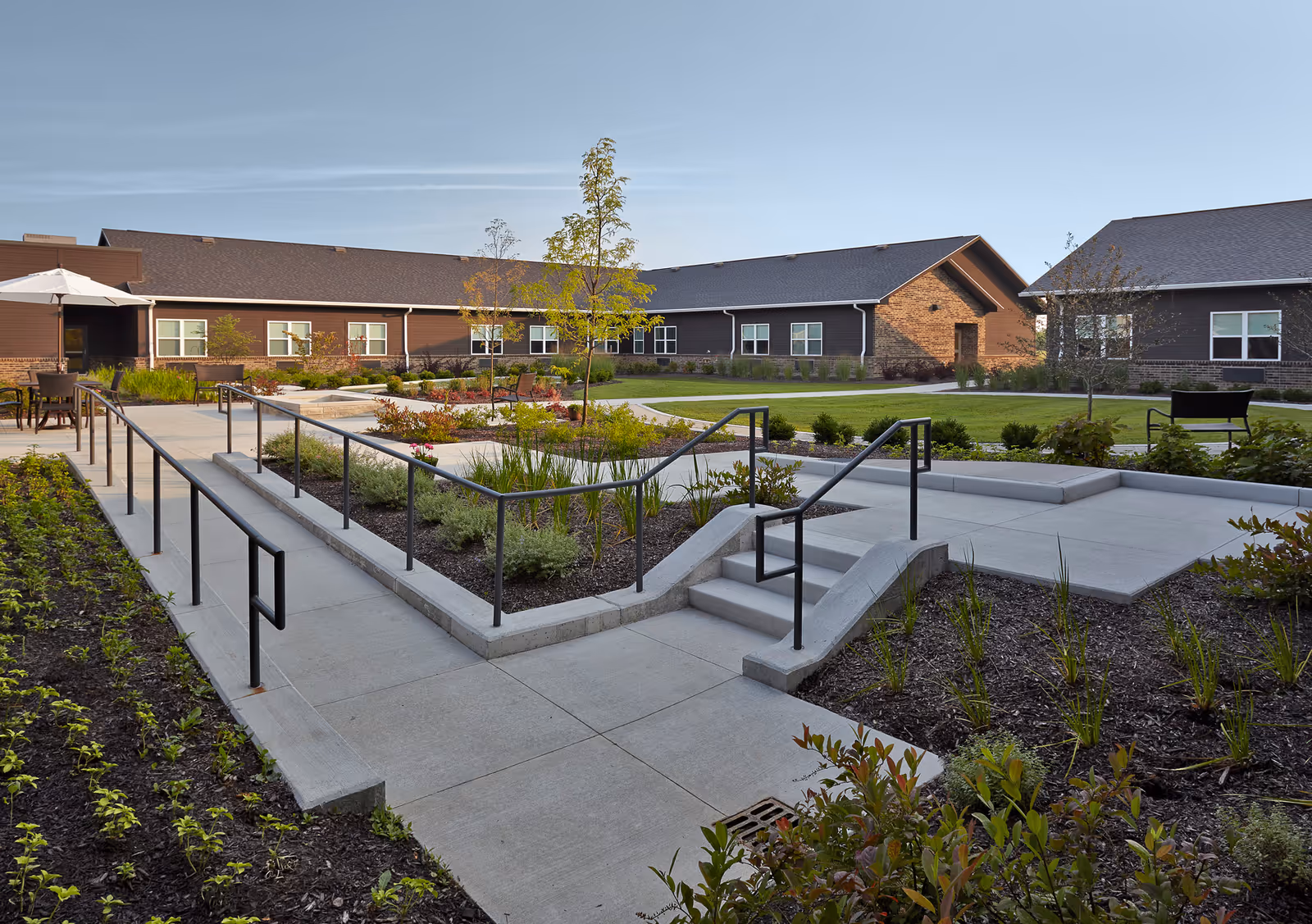 Outdoor courtyard area at Wellbrooke of Wabash featuring concrete walkways with handrails, landscaped garden beds, small trees, and a seating area with tables and chairs under an umbrella. The courtyard is surrounded by single-story buildings with brown siding and brick accents under a clear blue sky.