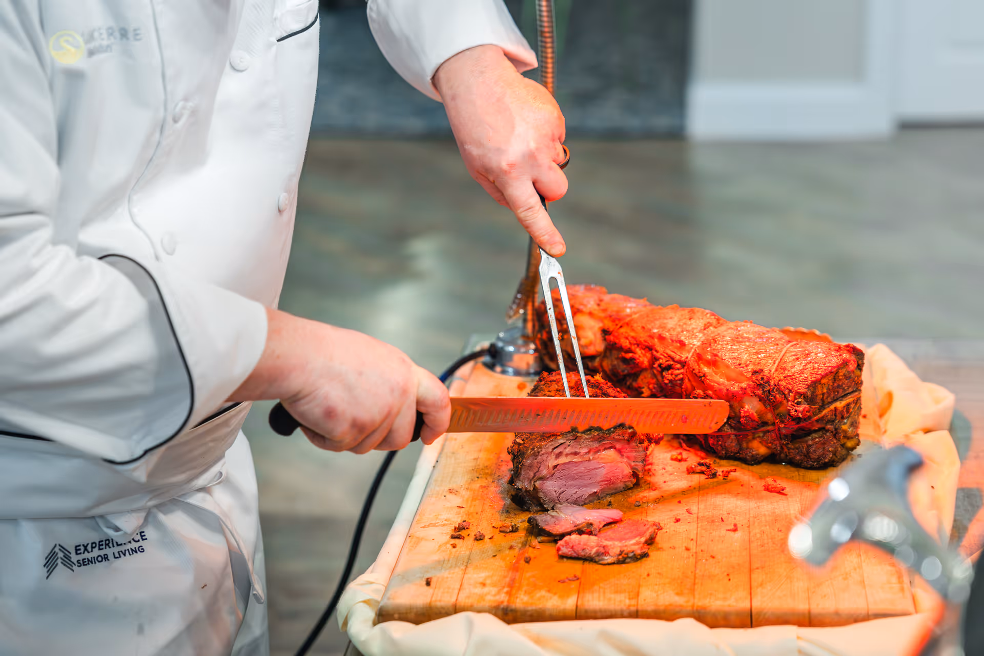 A chef wearing a white uniform is slicing a large piece of cooked roast beef on a wooden cutting board using a carving fork and knife.